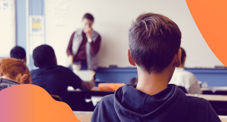 Frontalunterricht in eine Klasse: Im Vordergrund sieht man einen Schüler, der gerade im Unterricht sitzt. An der Tafel steht ein Lehrer.